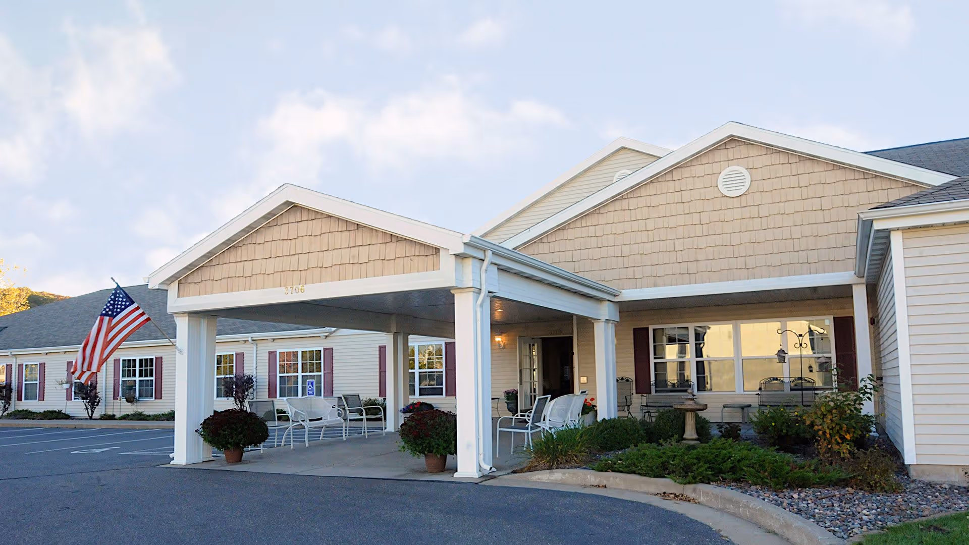 Front entrance of a single-story senior living facility with a covered porte-cochere, outdoor seating, an American flag, and landscaping.