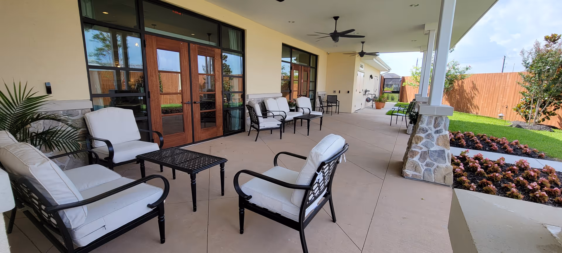 Covered outdoor patio area with cushioned metal chairs and small tables, ceiling fans, and a view of a garden with flowers and a wooden fence at The Harbor Assisted Living.