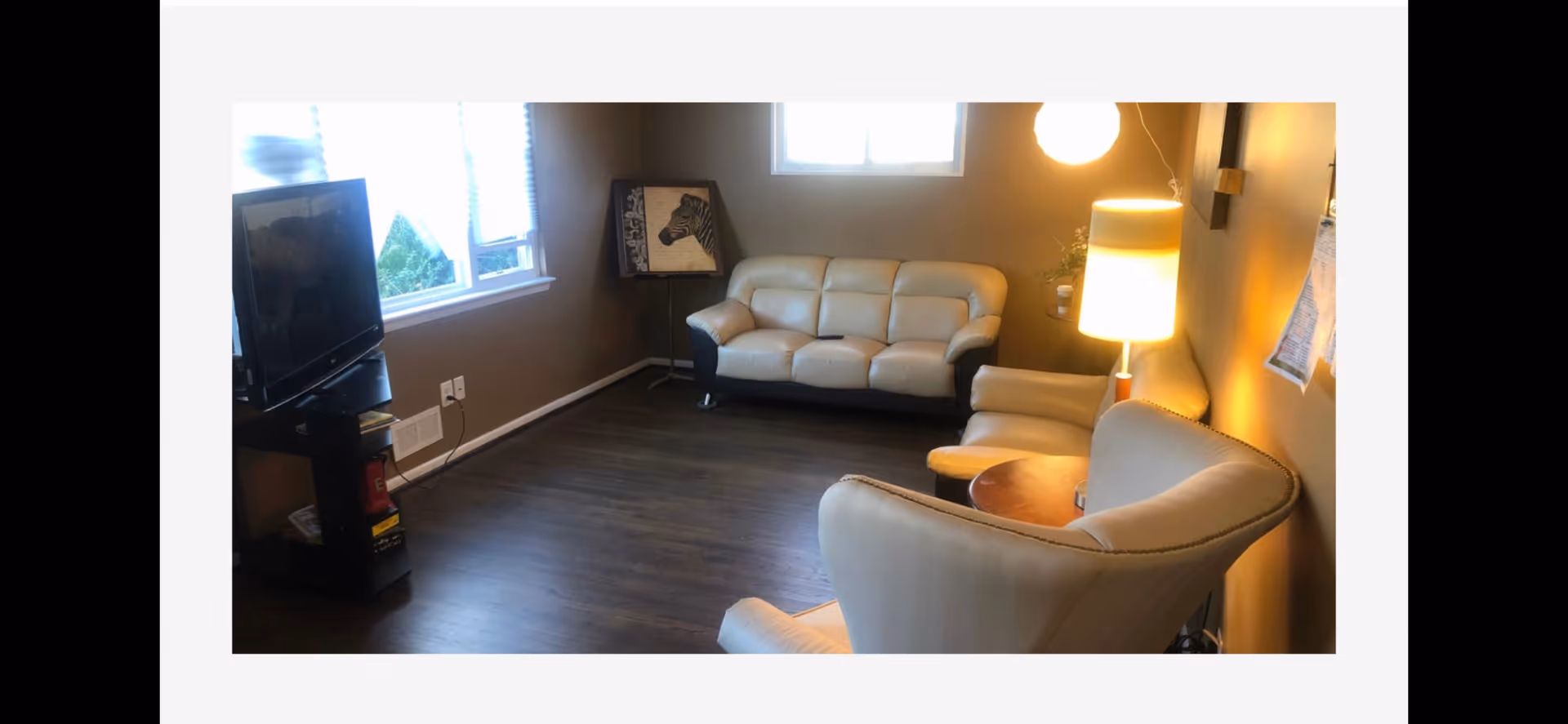 A cozy living room with a beige leather sofa and two matching armchairs arranged around a small wooden round table. A floor lamp with a white shade is turned on, casting warm light on the beige walls. A flat-screen TV is placed on a black stand near a window with white blinds. There is a framed zebra artwork resting on a stand in the corner.