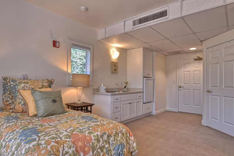 Studio-style bedroom with a floral bed, nightstand lamp, and a small built-in sink and cabinets.
