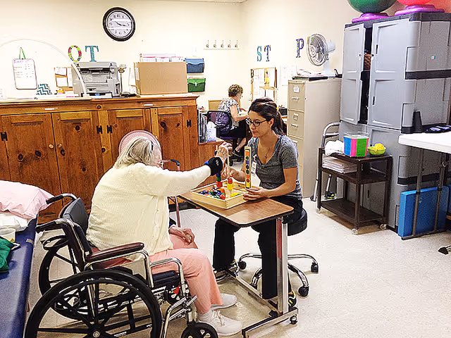 An elderly woman in a wheelchair playing a tabletop game with a younger woman seated across from her in a room with wooden cabinets, filing cabinets, and office supplies. Another person is seated at a desk in the background.