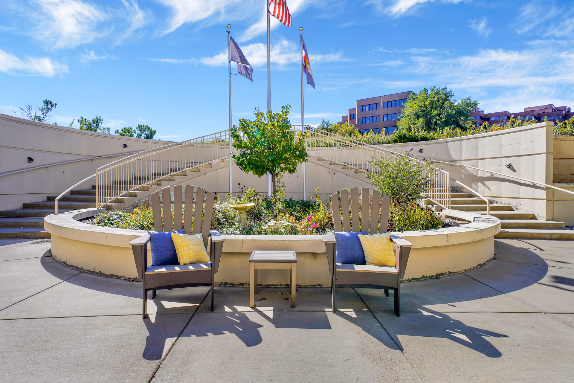 Outdoor seating area with two wooden chairs featuring blue and yellow cushions, a small table between them, and a circular raised garden bed with plants and flowers. Behind the garden bed are two staircases with railings leading up to a higher level. Three flagpoles with flags are visible in the background under a blue sky with some clouds.