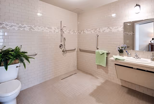 A spacious, accessible bathroom with beige tiled walls and floor. The bathroom features a walk-in shower with grab bars and a handheld showerhead. There is a white toilet with a green plant on top, a sink with a countertop, a mirror, and a green towel hanging on a grab bar. A small vase with flowers is placed on the sink counter.