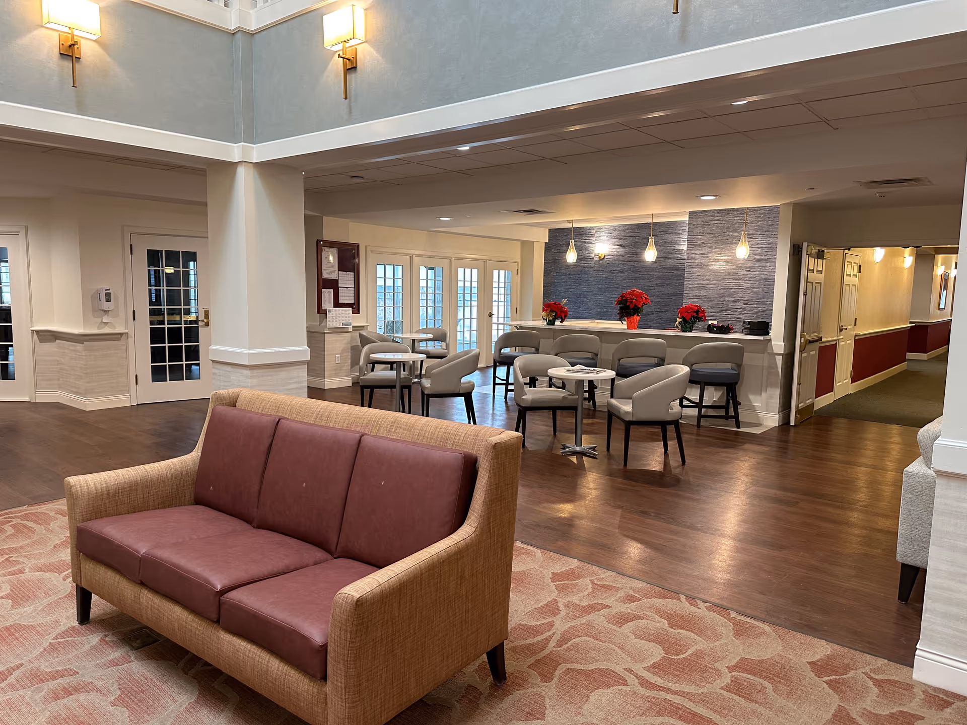 A spacious and well-lit common area in a senior living facility featuring a maroon cushioned sofa on a patterned carpet in the foreground. In the background, there are several small tables with chairs and a counter with bar stools, decorated with red poinsettia plants and pendant lights hanging from the ceiling. The walls are light-colored with some blue textured accent wall behind the counter. Doors and hallways lead to other parts of the facility.