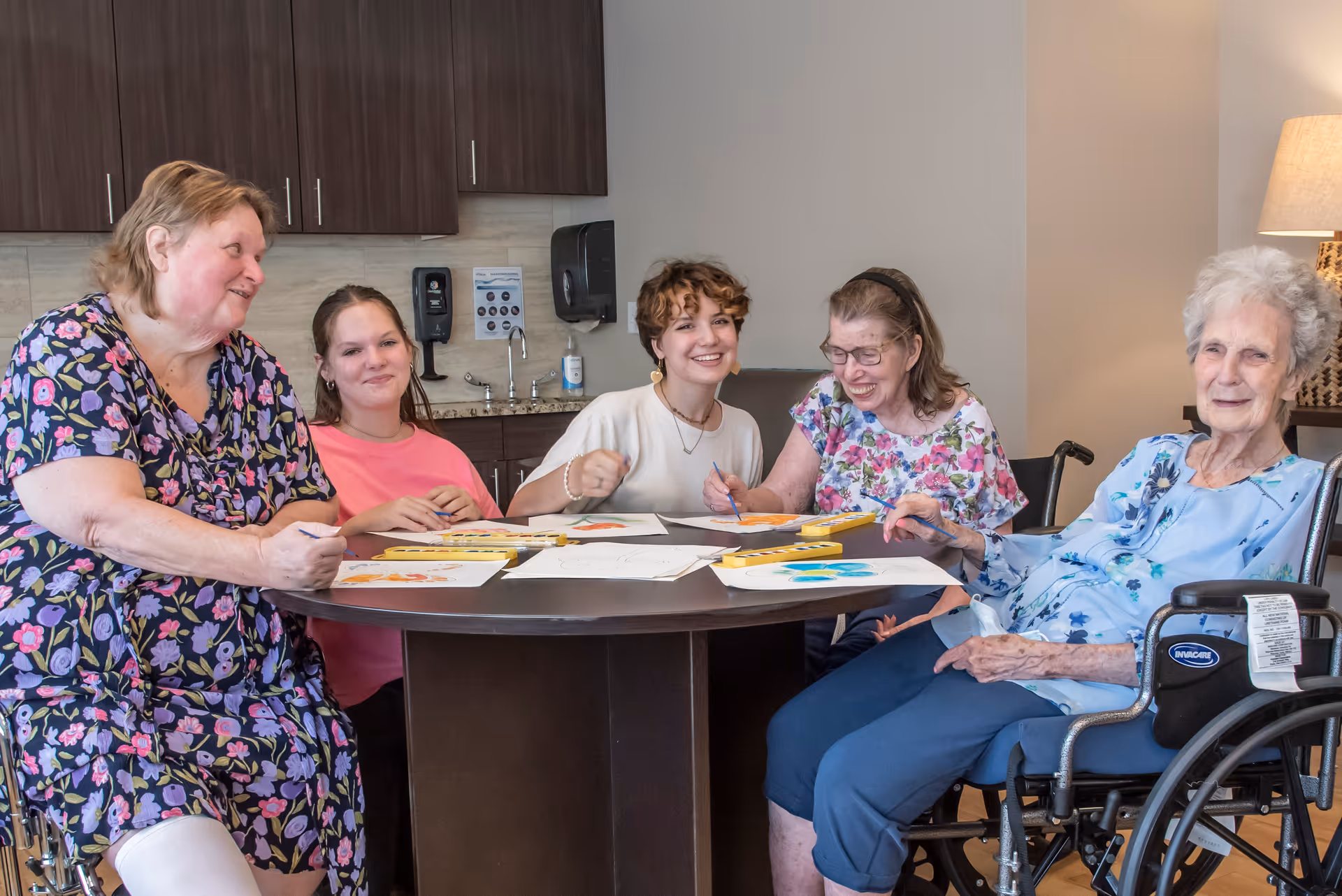 Five women sitting around a round table engaged in painting and drawing activities inside a room with cabinets and a sink in the background. Two of the women are in wheelchairs, and all are smiling and enjoying the activity.