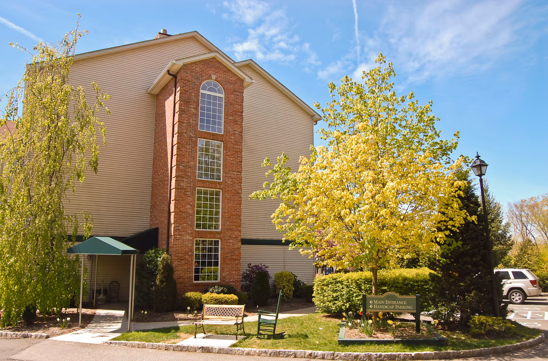 Exterior view of a multi-story assisted living facility building with beige siding and a central brick section featuring tall windows. The scene includes a green canopy over an entrance, a bench, a rocking chair, trees with green and yellow leaves, a lamp post, and a sign indicating the main entrance and handicap parking. A white SUV is parked in the background under a clear blue sky.