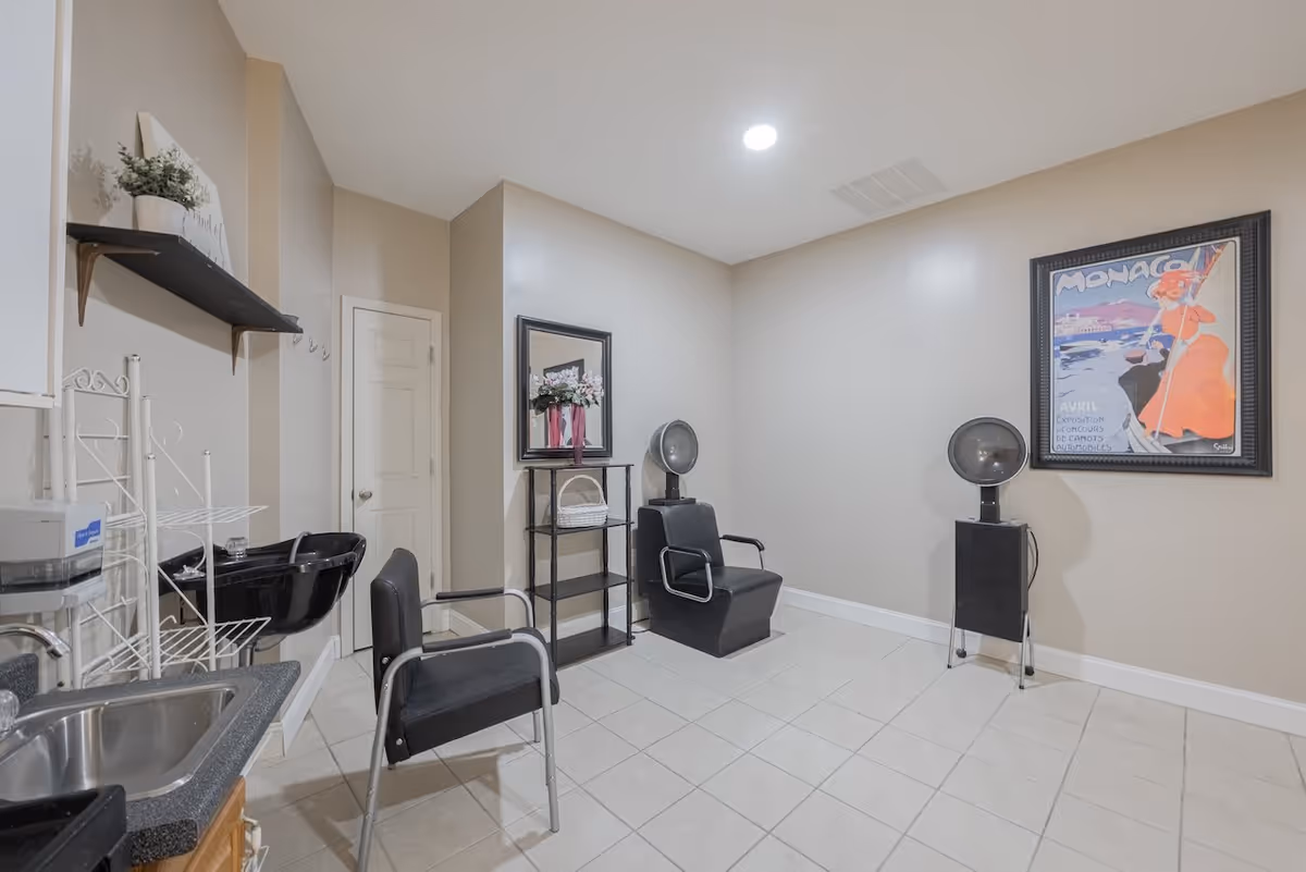 Interior of a senior living facility room with two black salon chairs, a black hair washing sink, a small black shelving unit with a mirror and flowers, a framed Monaco poster on the wall, and a tiled floor.