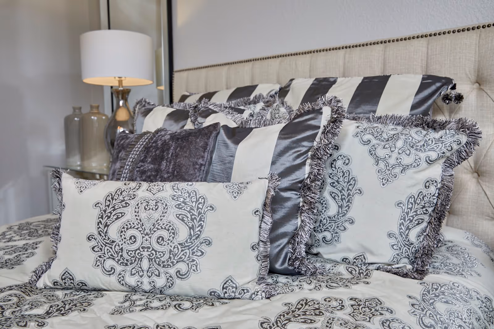 Close-up view of a bed with decorative pillows in various patterns including striped and floral designs, a beige upholstered headboard with nailhead trim, and a bedside table with a lamp and two glass vases.