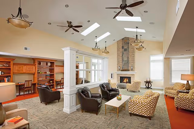 A spacious and well-lit living room area with a high ceiling featuring skylights and ceiling fans. The room includes a stone fireplace, multiple comfortable chairs with patterned upholstery, leather armchairs, a coffee table, and built-in wooden desks with chairs along one wall. The space is decorated in warm tones with beige walls and carpeted floors.