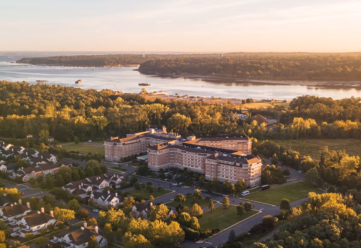 Aerial view of The Harborside senior living facility surrounded by trees and residential houses, with a large body of water and forested land in the background during sunset.