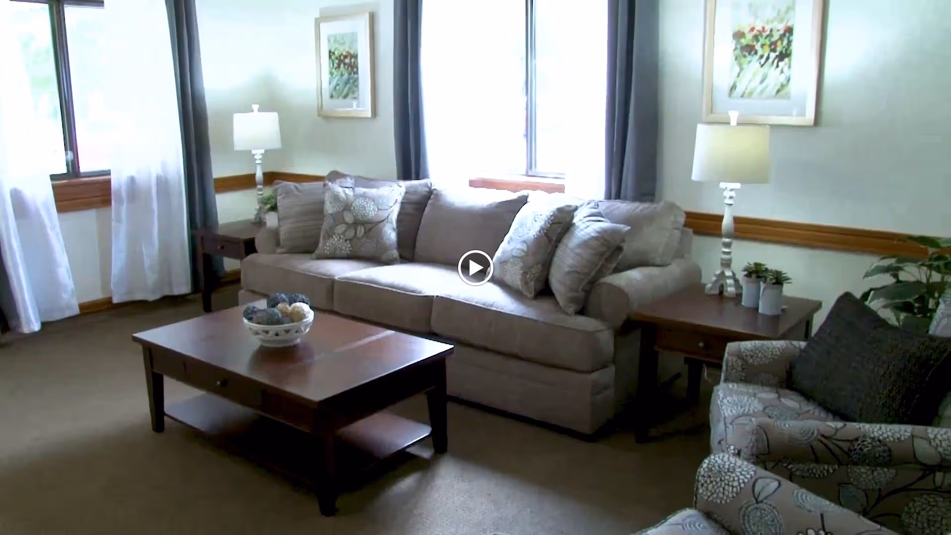A cozy living room with a beige sofa adorned with patterned cushions, a wooden coffee table with a decorative bowl, two side tables each with a lamp, framed artwork on the walls, and large windows with curtains letting in natural light.