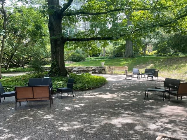 Outdoor patio area with several sets of black metal chairs and benches arranged on a gravel surface under large trees providing shade. There is a low stone wall and green grass in the background, creating a peaceful garden setting.