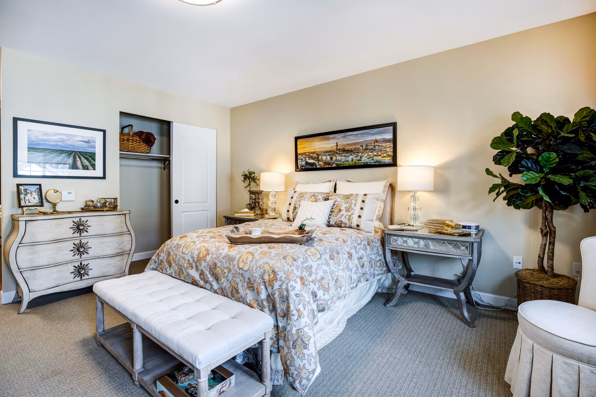 A cozy bedroom with a patterned bedspread on a bed, two bedside tables with lamps, a cushioned bench at the foot of the bed, a decorative chest of drawers, a potted plant, and framed artwork on the walls.