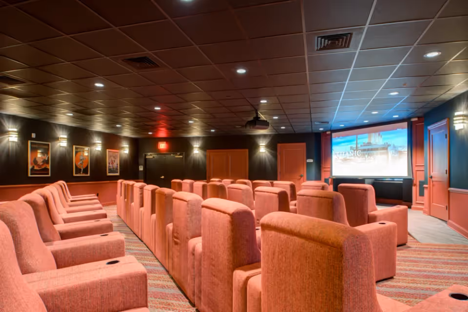 Interior view of a small theater room with rows of comfortable pink upholstered recliner chairs facing a large screen displaying the movie Titanic. The room has dark walls with framed posters, soft wall lighting, and a ceiling-mounted projector.
