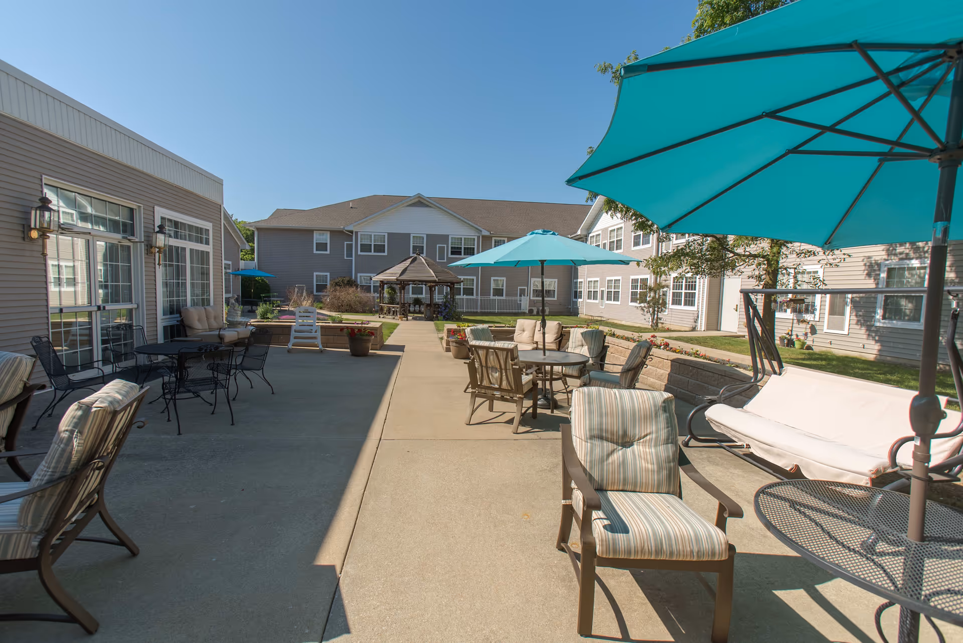 Outdoor patio area at Eagle Ridge of Decatur featuring multiple seating arrangements with cushioned chairs, tables with teal umbrellas, and a swing bench. The patio is surrounded by a two-story building with large windows and a gazebo in the background under a clear blue sky.