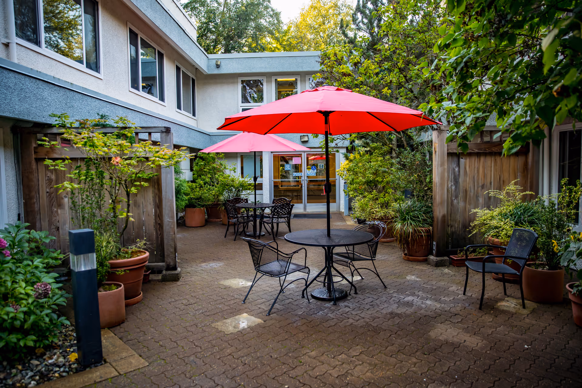 Outdoor courtyard area with brick paving, featuring black metal tables and chairs under red umbrellas. The space is surrounded by potted plants and greenery, with a two-story building in the background and glass doors leading inside.