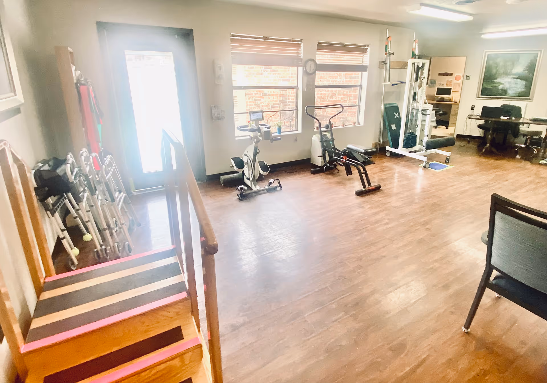 A spacious room with wooden flooring featuring exercise equipment including a stationary bike, rowing machine, and a multi-functional gym machine. There is a small set of wooden stairs with handrails on the left side, several walkers lined up against the wall, and two large windows letting in natural light. In the background, there is a desk with a computer and a framed painting on the wall.