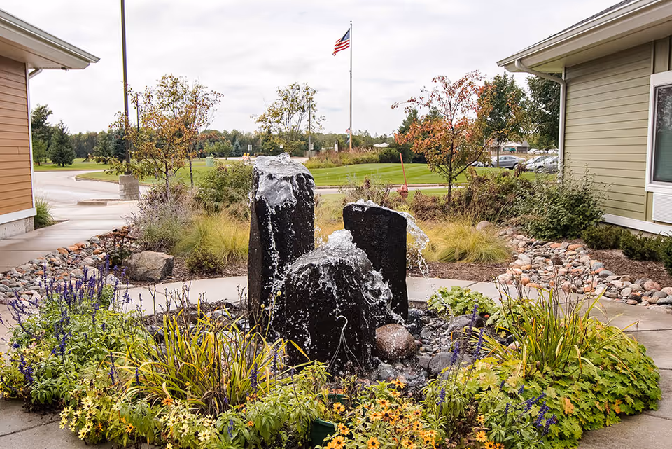 Outdoor garden area at Edgewood Blaine featuring a water fountain with three large black stone pillars surrounded by colorful flowers and greenery. In the background, there is an American flag on a flagpole, trees, and a parking area with cars.