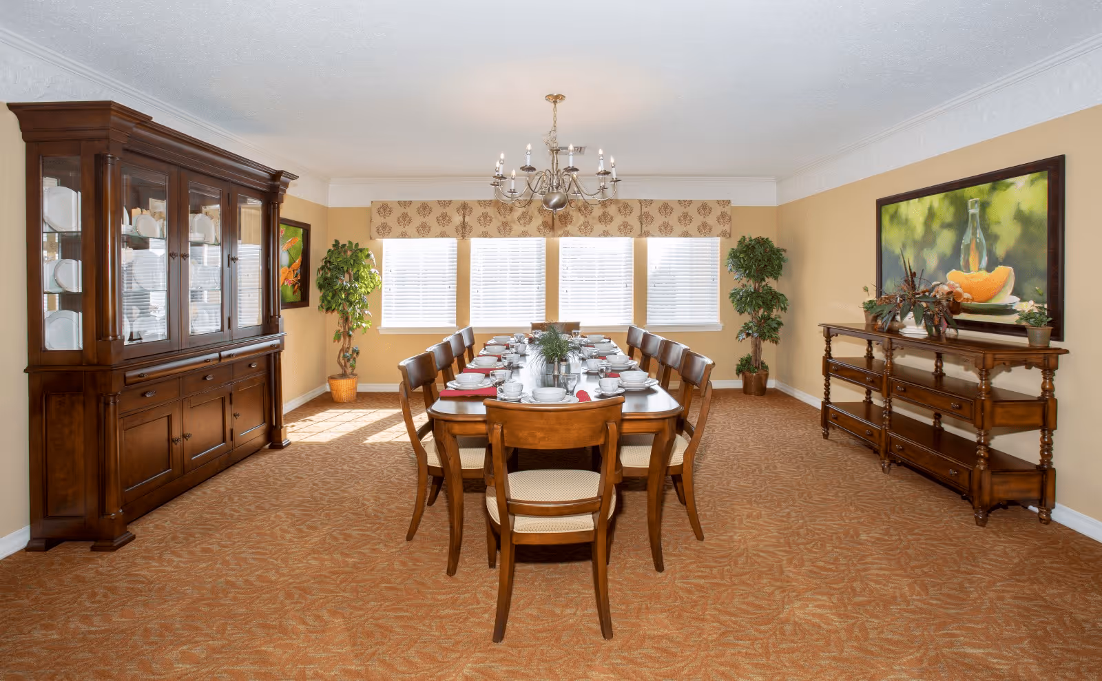 A formal dining room with a long wooden dining table set for a meal with plates, bowls, cups, and napkins. The room features a large wooden china cabinet on the left, a wooden sideboard on the right, two potted plants near the windows, and a chandelier hanging from the ceiling. The walls are painted beige, and there is a large painting above the sideboard.