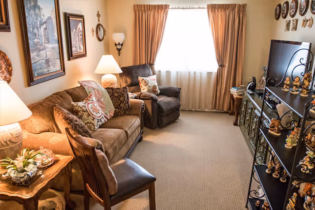 A cozy living room with a brown sofa adorned with patterned pillows, a dark brown recliner chair, a wooden side table with a lamp, and a wooden chair. The room has beige carpet, light-colored walls with framed artwork and decorative crosses, a window with beige curtains, and a black shelving unit displaying numerous small figurines. A TV is placed on a cabinet opposite the seating area.