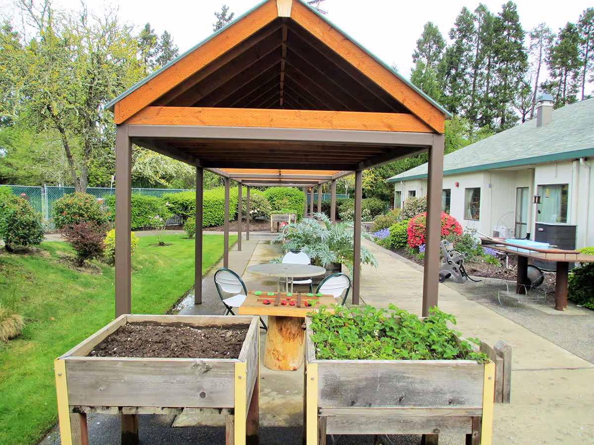 Covered outdoor walkway and garden area with raised planters, tables and chairs next to the facility building.