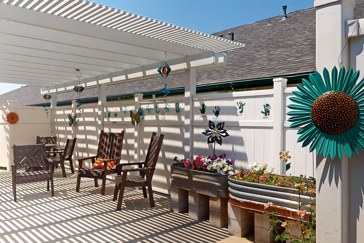Outdoor patio area with wooden chairs and a table under a white pergola casting striped shadows. The white fence behind is decorated with colorful handprint art, butterfly and flower ornaments. There are raised garden beds with blooming flowers and a large green sunflower decoration on the right side.