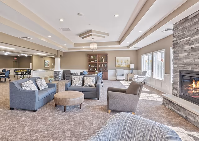 A spacious and well-lit living room area in an assisted living facility featuring multiple gray upholstered sofas and armchairs arranged around a round ottoman. There is a stone fireplace with a fire burning on the right side, large windows letting in natural light, and a wooden bookshelf with decorative items against the far wall. The room has a beige carpet and neutral-colored walls with white trim.