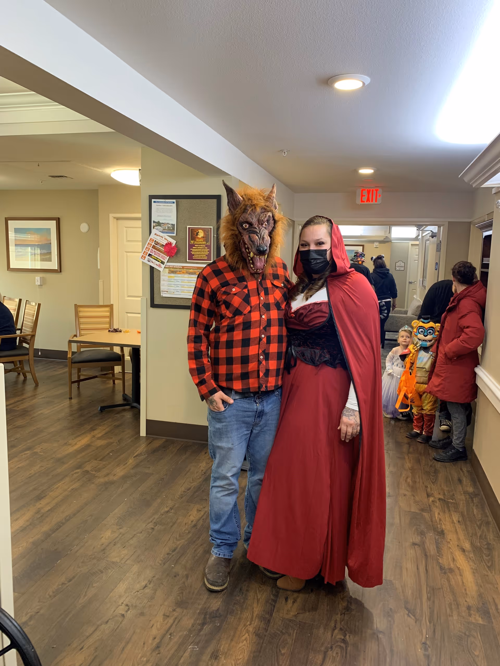 Two adults dressed in Halloween costumes stand in a hallway of a senior living facility. One person is wearing a werewolf mask with a red and black checkered shirt and jeans. The other person is dressed as Little Red Riding Hood with a red cape and a black face mask. In the background, children in costumes and other adults are visible near a seating area with wooden chairs and tables. The hallway has wood flooring and neutral-colored walls with framed pictures and a bulletin board.