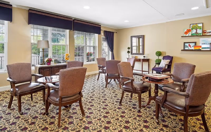Sunlit communal lounge with multiple small tables and upholstered chairs arranged on a patterned carpet near large windows.