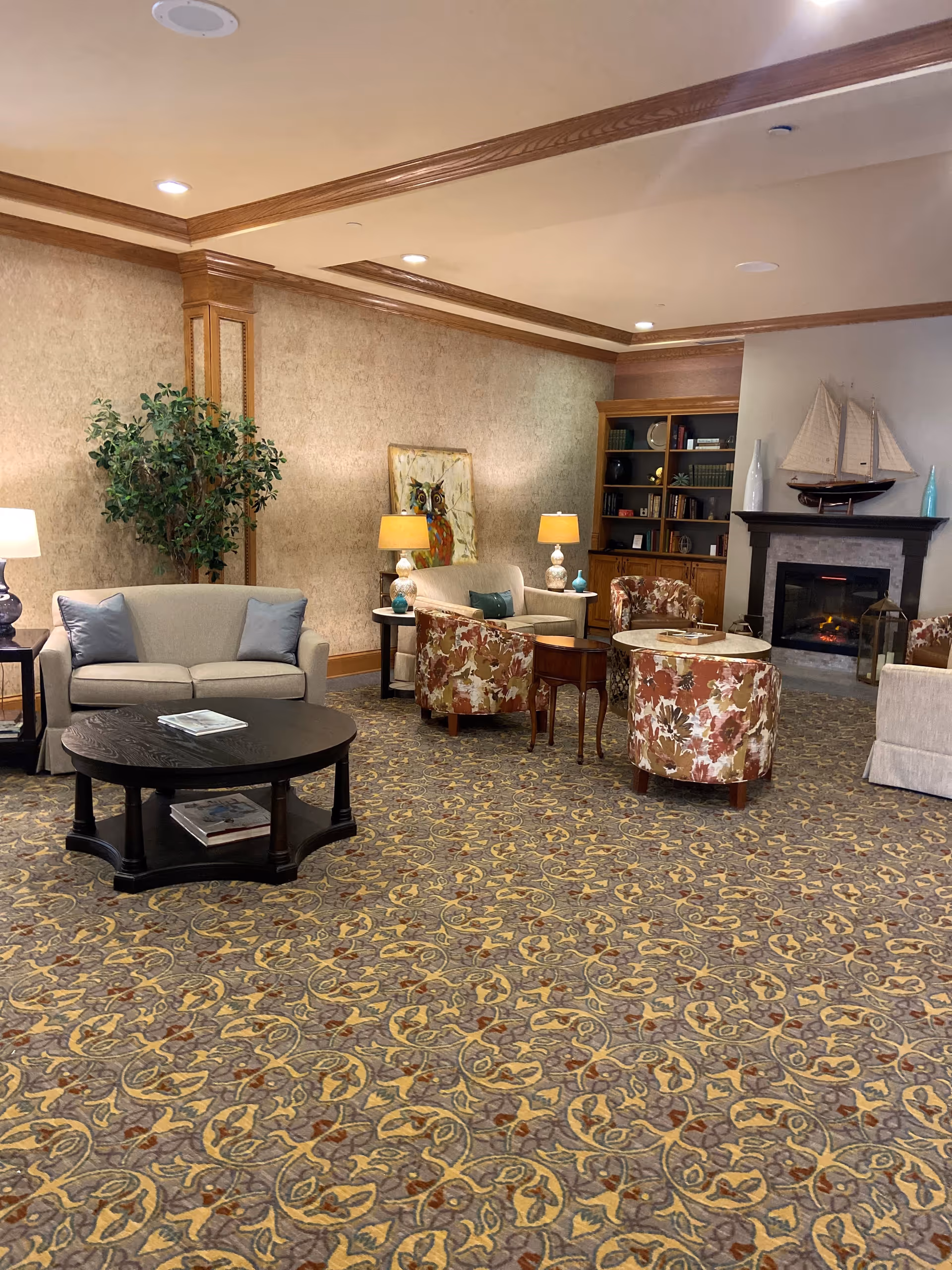 A cozy living room area with patterned carpet, beige and floral upholstered chairs, a beige loveseat with blue pillows, a round black coffee table, side tables with lamps, a bookshelf, and a fireplace with a decorative sailboat on the mantel.