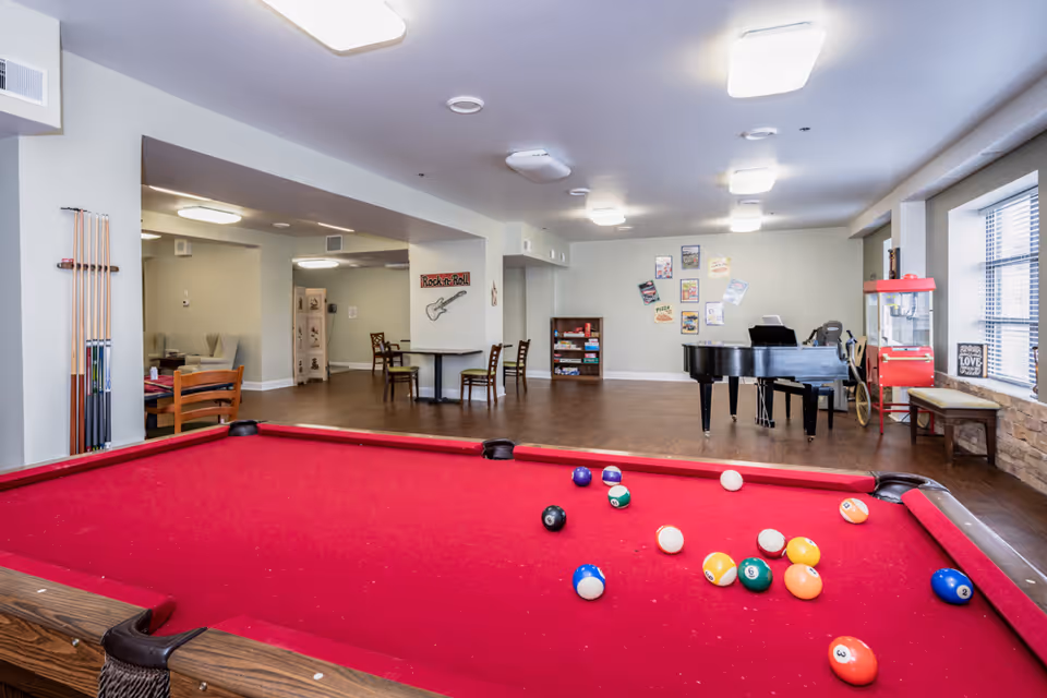 A recreational room with a red pool table in the foreground, several pool cues mounted on the wall to the left, a black grand piano, a popcorn machine, and a few tables and chairs scattered around. The walls are decorated with various posters and signs, and there is a window with blinds on the right side.