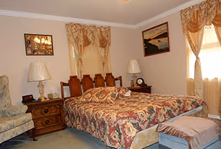 A neatly furnished bedroom with a floral bedspread, two bedside tables with lamps, an armchair, and curtained windows.