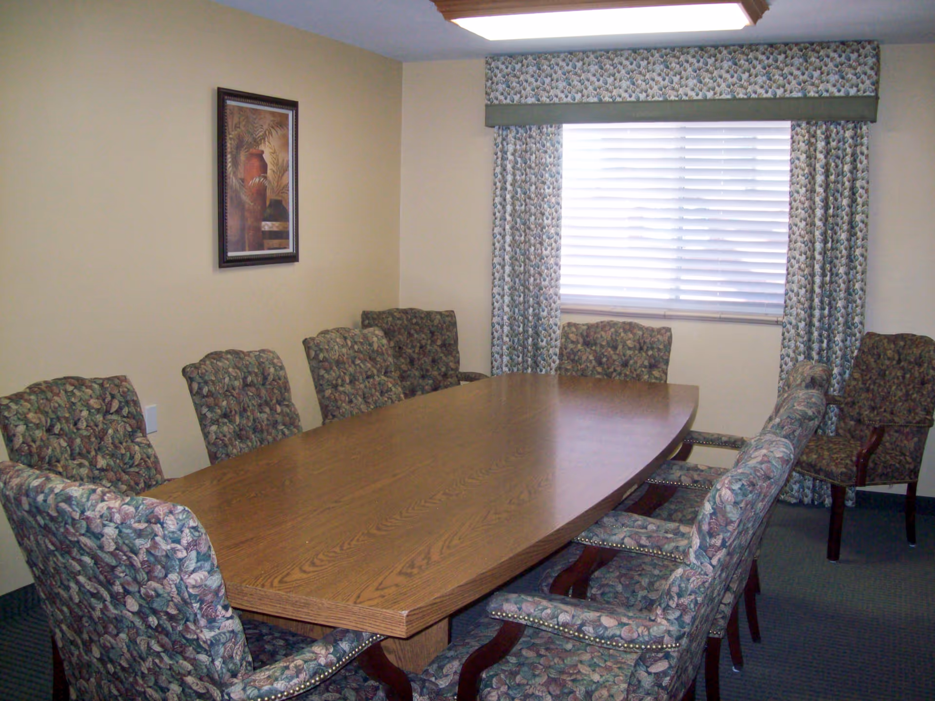 A conference or meeting room with a large wooden table surrounded by eight upholstered chairs with floral patterned fabric. There is a window with closed blinds and matching floral curtains. A framed picture hangs on the beige wall.