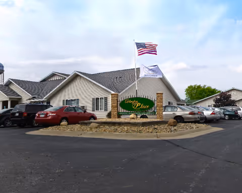 Parking lot in front of a single-story building with beige siding and a gray roof. In the center, there is a landscaped island with rocks and a sign that reads 'Arlington Place' along with two flagpoles flying the American flag and another flag. Several cars are parked around the building under a partly cloudy sky.