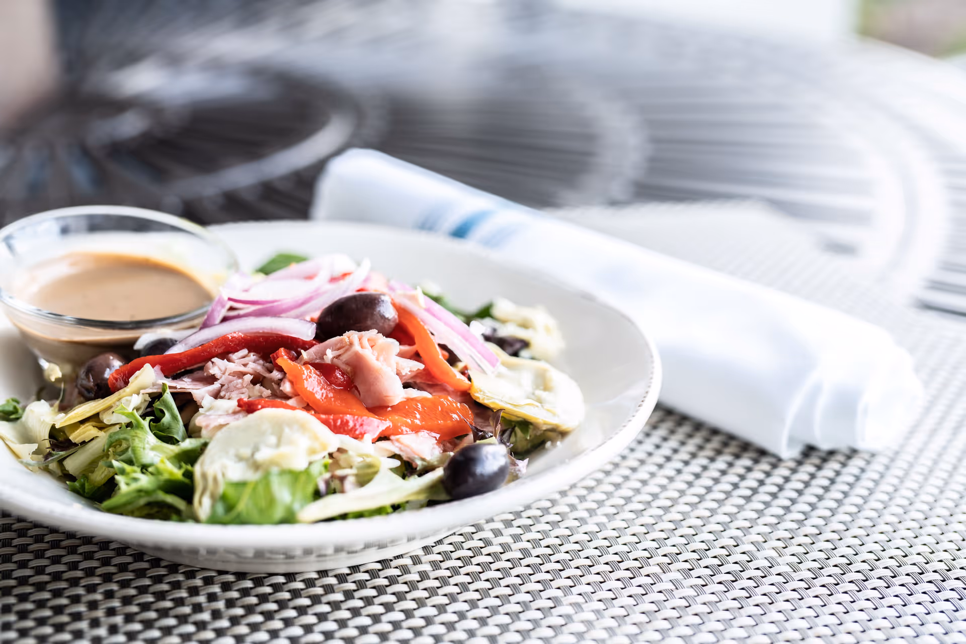 A fresh salad with mixed greens, sliced red onions, olives, roasted red peppers, artichoke hearts, and shredded meat served in a white bowl with a small glass container of dressing on the side. A rolled white napkin with a blue stripe is placed next to the bowl on a woven placemat.