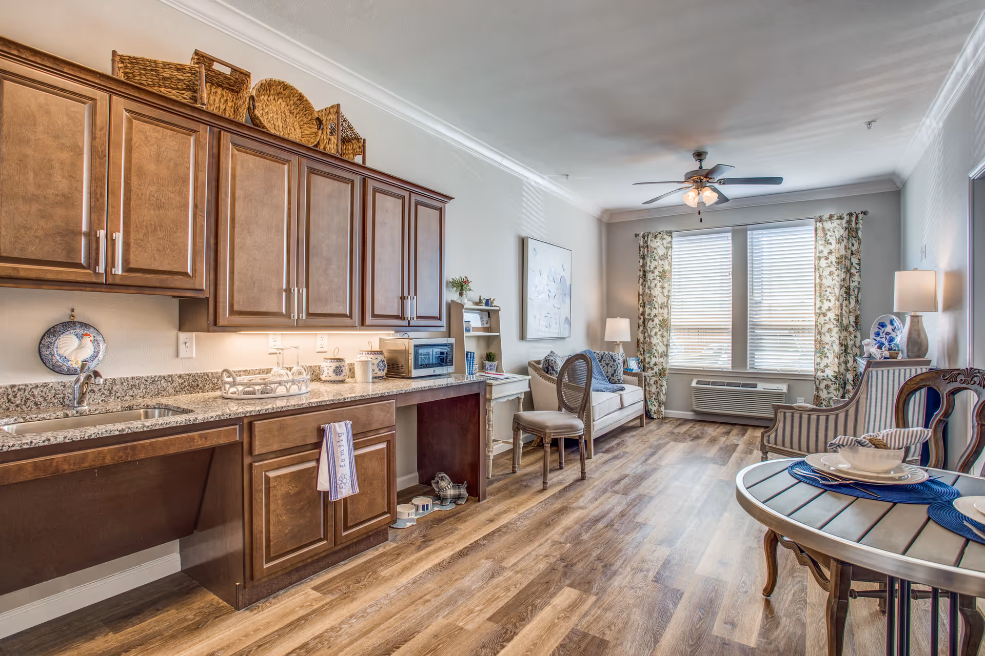 A bright and cozy living area with wooden flooring, a ceiling fan, and large windows with floral curtains. The room features a small kitchen area with brown cabinets and granite countertops on the left, a white sofa with cushions, a wooden chair, and a round dining table set with plates and bowls on the right.