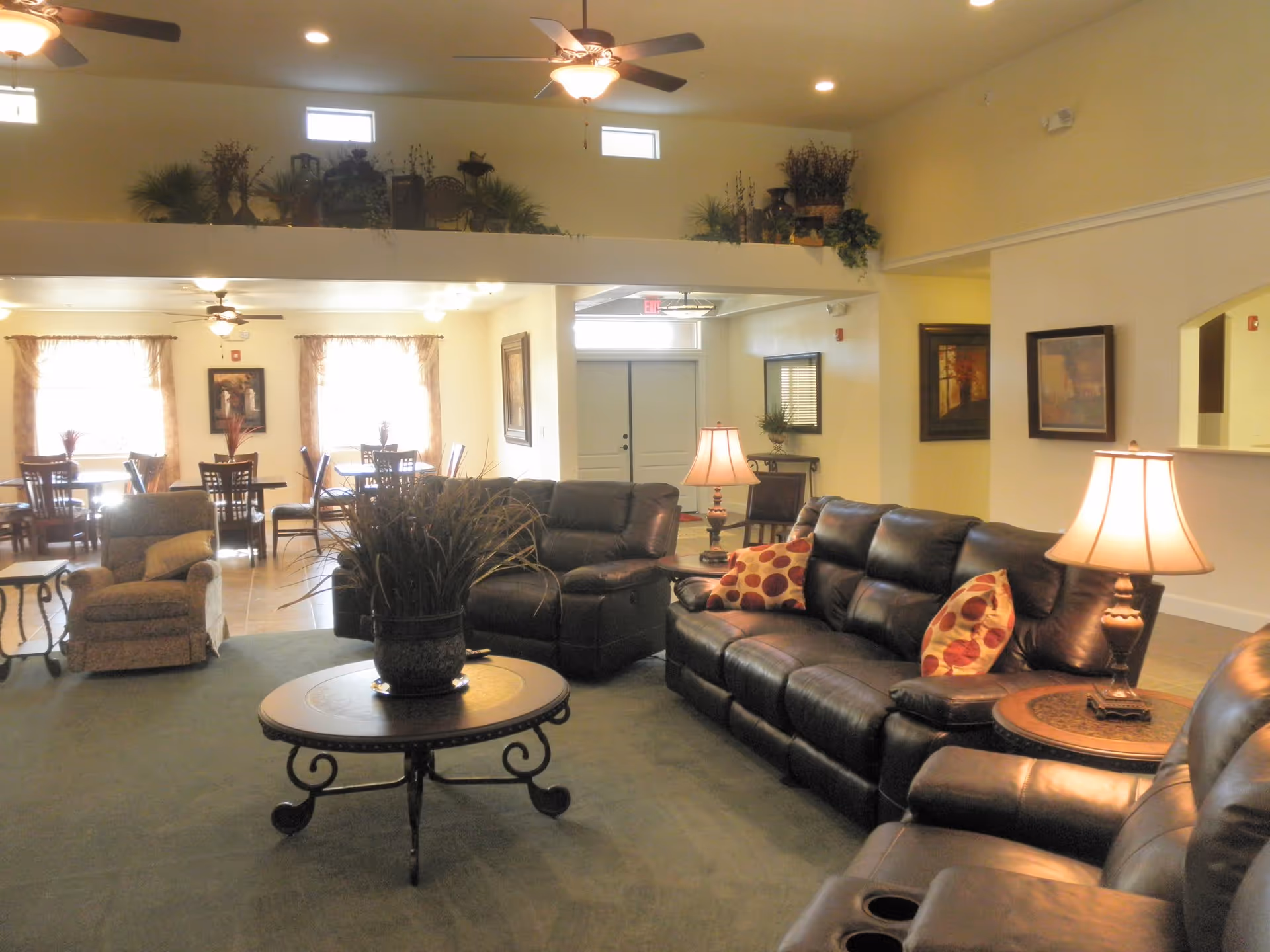 A cozy living room area in an assisted living facility with dark leather sofas adorned with patterned pillows, a round wooden coffee table with a potted plant, and two table lamps on side tables. In the background, there is a dining area with several tables and chairs near windows with curtains. The room has ceiling fans, recessed lighting, and decorative plants on a ledge above the living space.