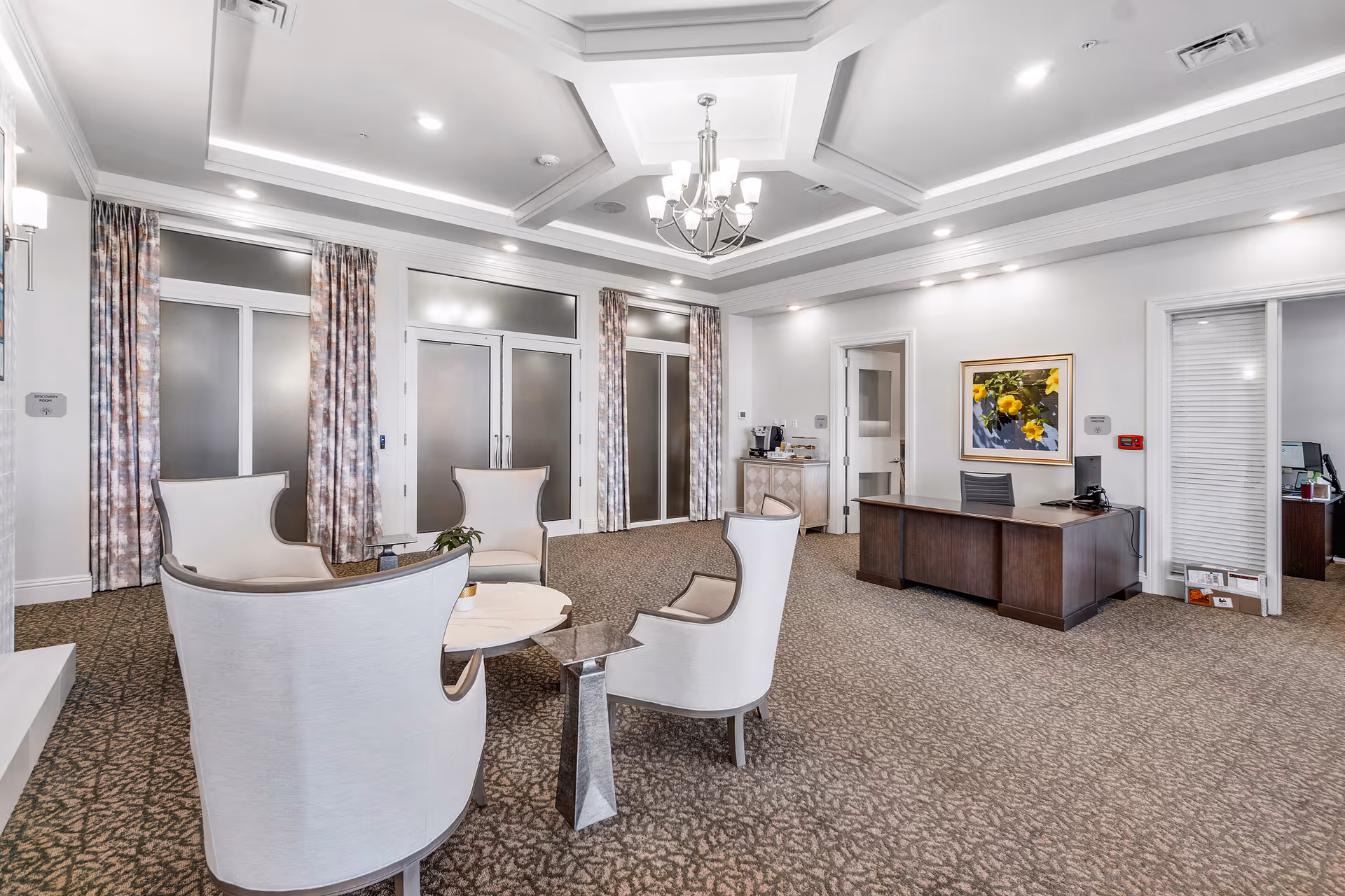 Well-lit reception lounge with upholstered armchairs around a small table, a decorative coffered ceiling with chandelier, and a wooden reception desk.