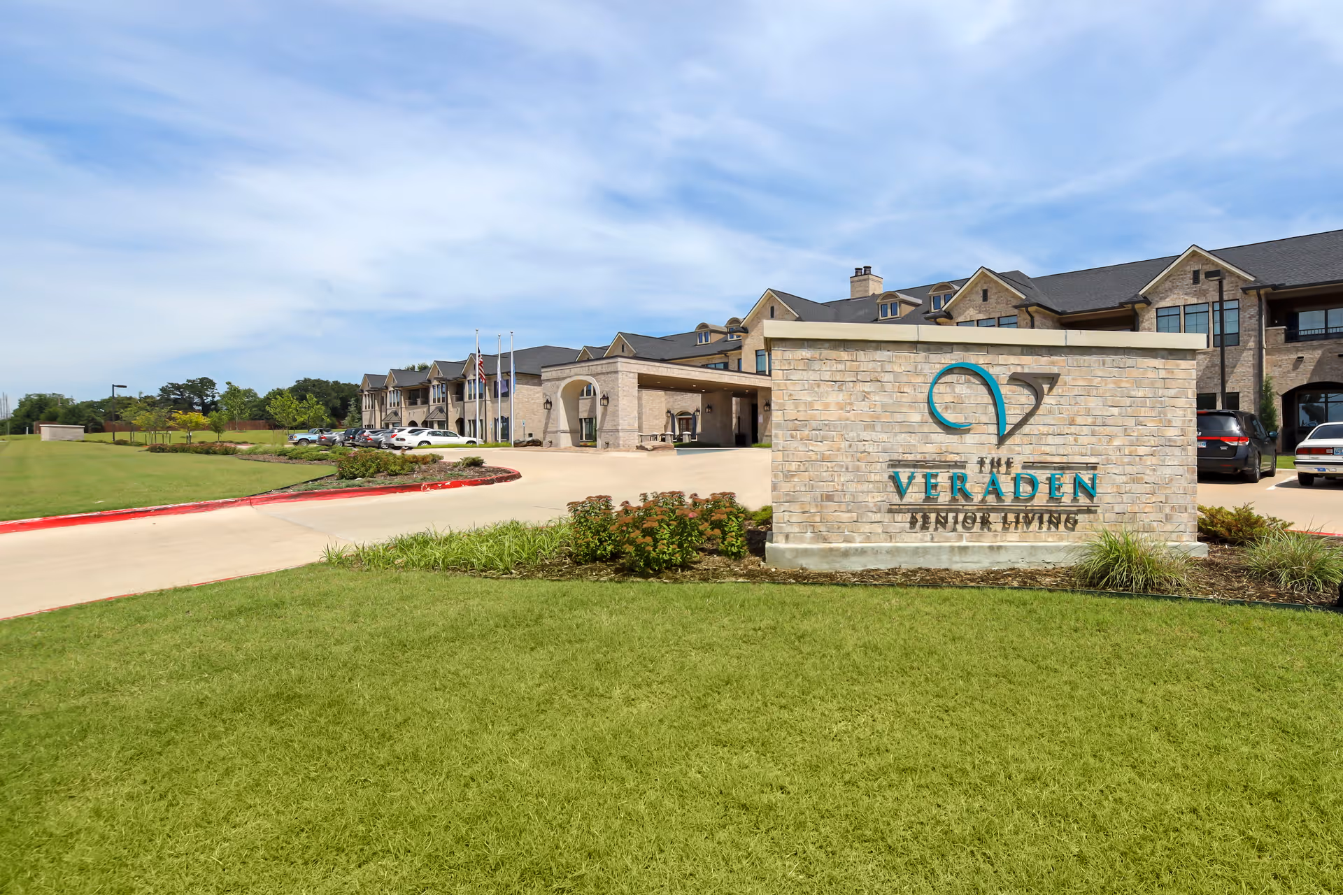 Exterior view of The Veraden Senior Living facility showing a large brick sign with the facility's name in front of a well-maintained lawn and driveway. The building is a multi-story structure with a stone facade, multiple windows, and a covered entrance. Several cars are parked near the entrance under a partly cloudy sky.