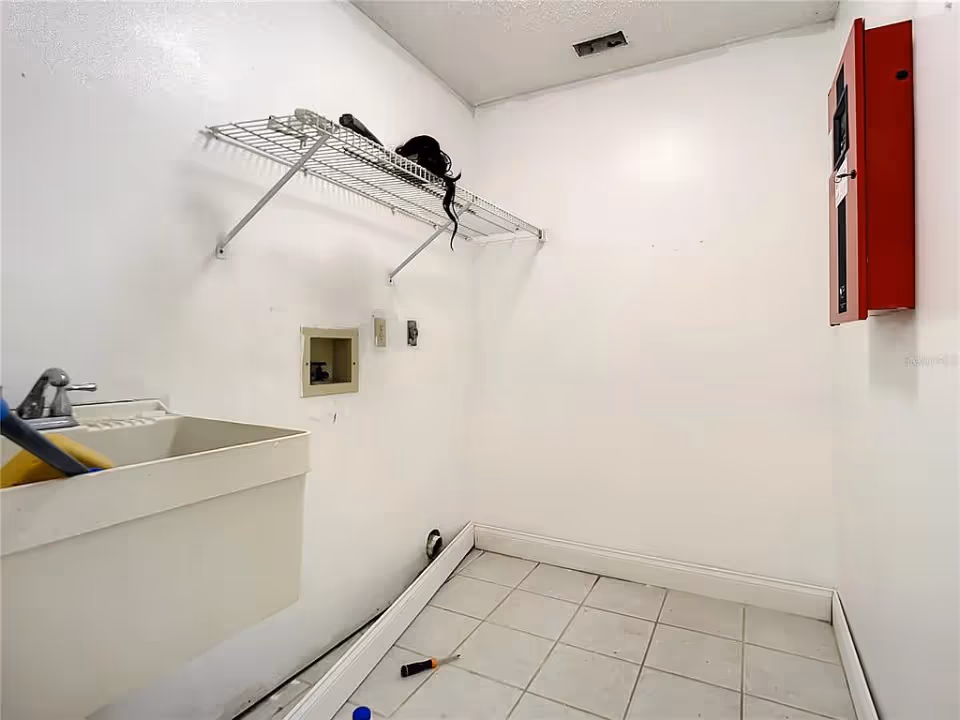 A small utility or laundry room with white tiled floor and white walls. There is a utility sink with a faucet on the left side, a wire shelf mounted on the wall above with some items on it, and a red electrical panel mounted on the right wall. The room appears empty otherwise, with some small objects on the floor.