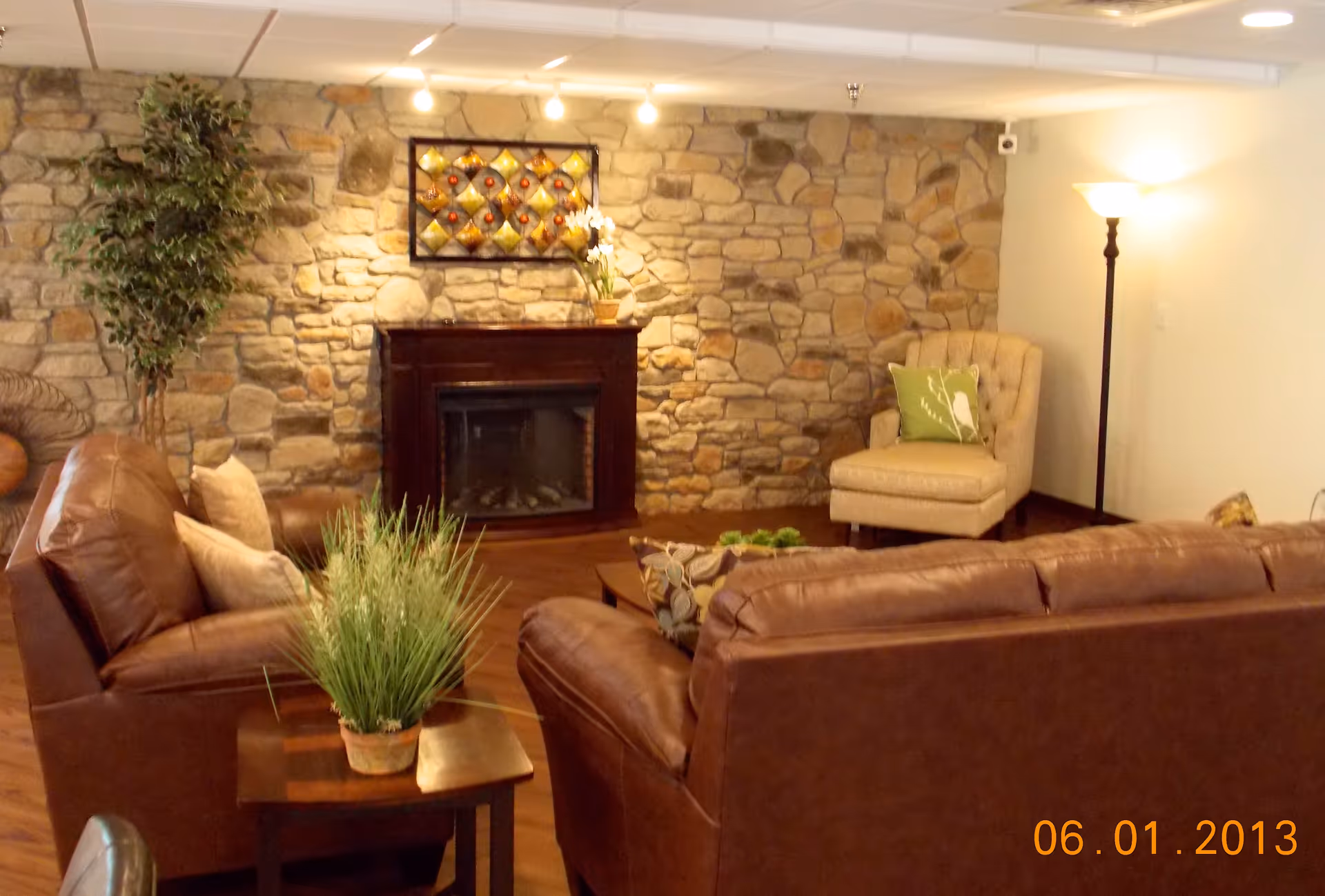 Cozy living room area with brown leather sofas, a stone wall with a dark wood fireplace, a beige armchair with a green pillow, a tall floor lamp, and decorative plants on a wooden side table and in the corner.