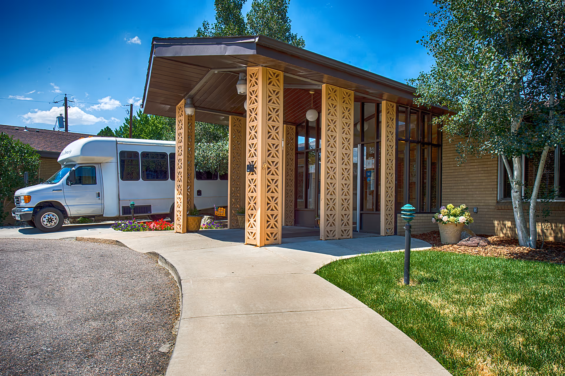 Entrance of a senior living facility with a covered walkway supported by decorative columns. A white shuttle bus is parked nearby, and there are trees, green grass, and flower pots around the entrance under a clear blue sky.
