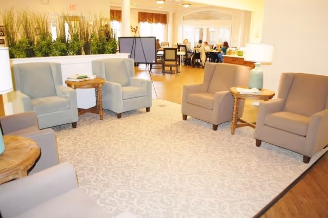A bright and spacious common area with several upholstered armchairs arranged around small wooden side tables with lamps. In the background, there is a dining area with people seated at tables. The room features light-colored walls, large windows with curtains, and a patterned carpet on the floor.