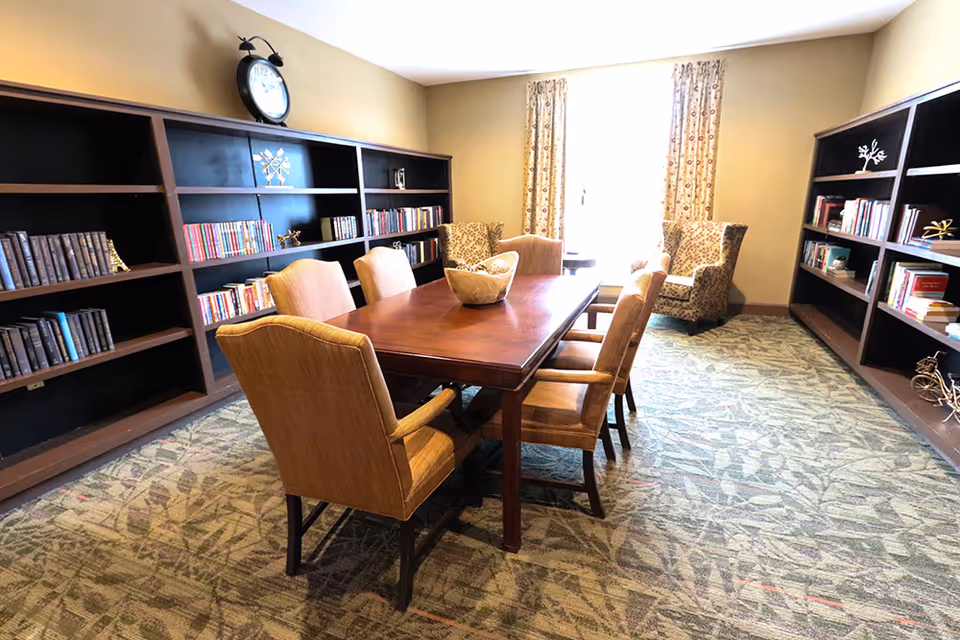 A communal library/reading room with a long wooden table surrounded by upholstered chairs and bookshelves along the walls.