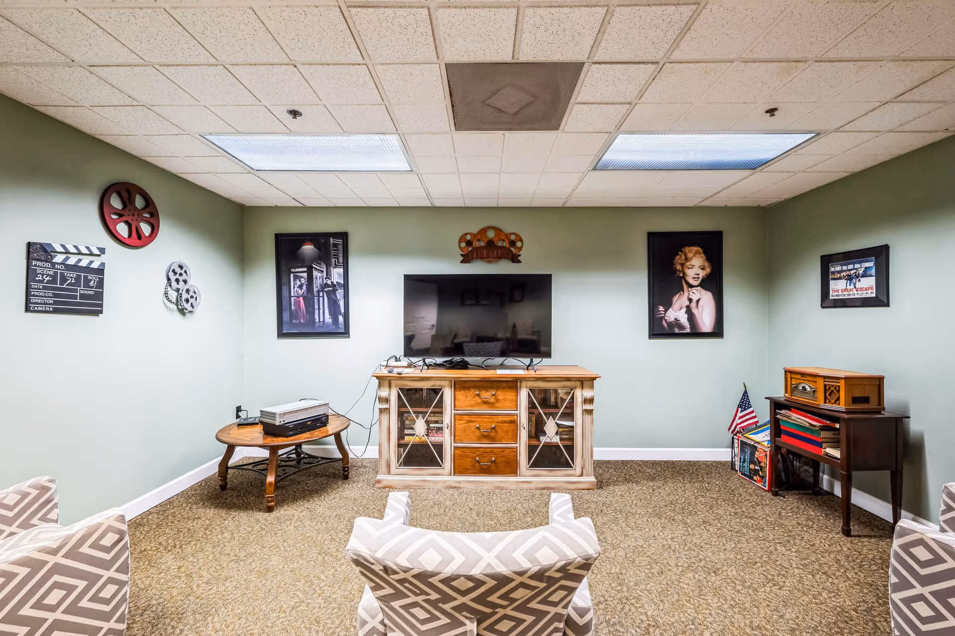 A cozy living room with light green walls and a beige carpet. The room features a flat-screen TV on a wooden cabinet with glass doors, a small round wooden table with electronic equipment, and a vintage-style record player on a small wooden table. The walls are decorated with framed pictures, including a black and white photo, a portrait of Marilyn Monroe, and movie-themed decor such as a film reel and a clapperboard. There are patterned armchairs facing the TV.