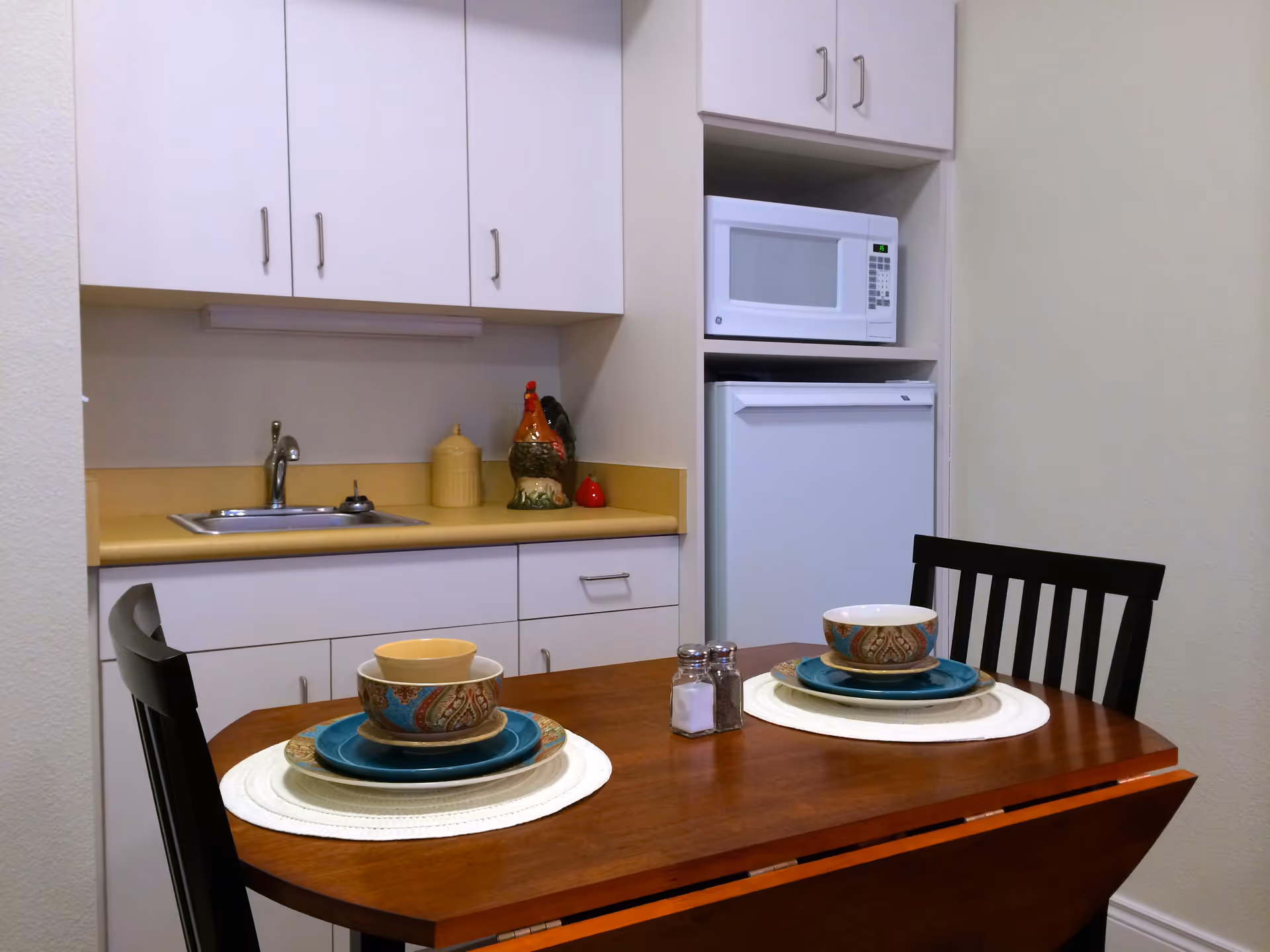 Small kitchen nook with a sink, microwave and mini-fridge and a wooden table set for two.