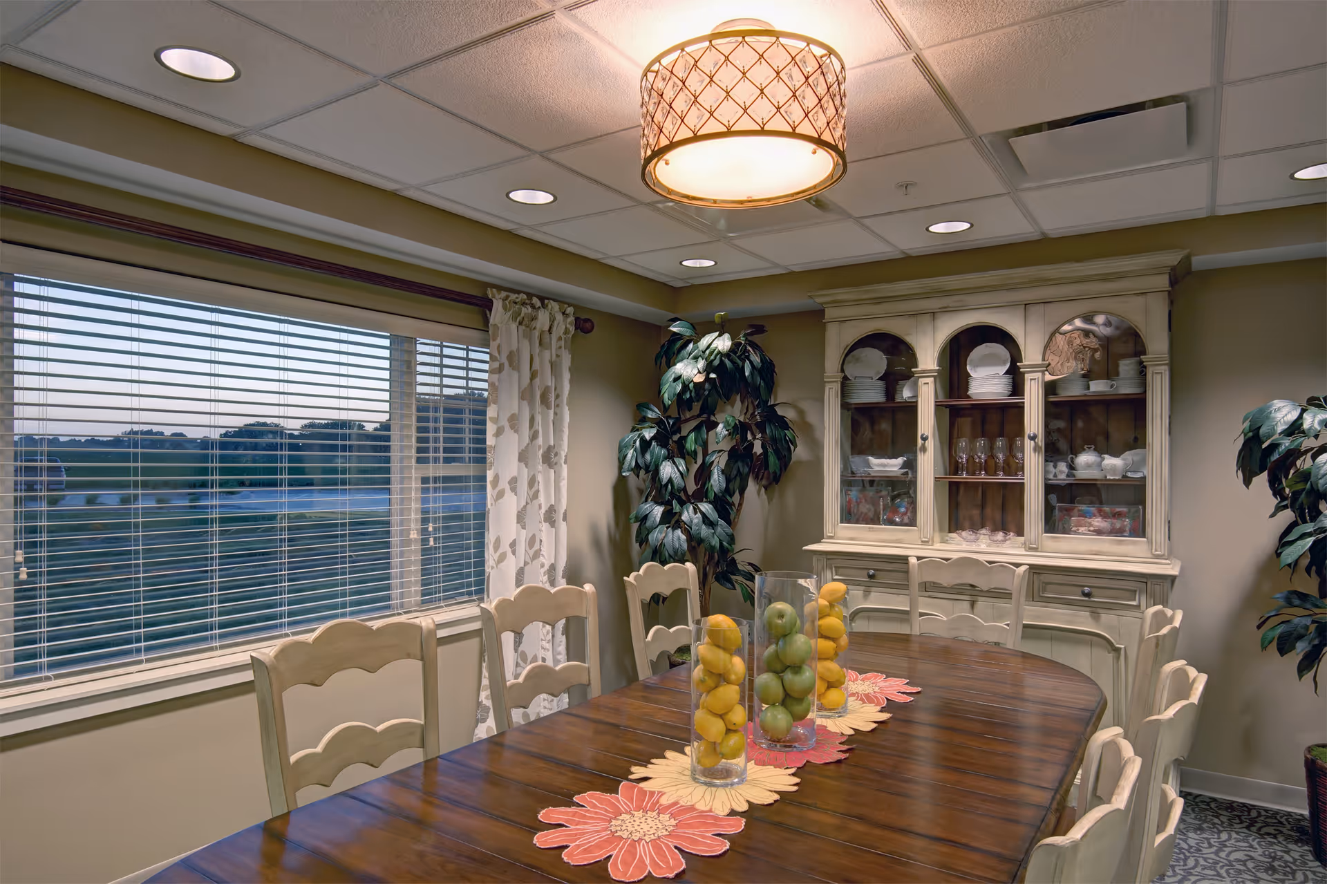 A dining room with a long wooden table surrounded by cream-colored chairs. The table has three glass vases filled with green and yellow apples, placed on floral placemats. There is a large window with blinds and floral curtains on the left wall. A cream-colored cabinet with glass doors displaying plates and glassware is against the back wall. Two large potted plants are positioned on either side of the cabinet. A decorative ceiling light fixture hangs above the table.