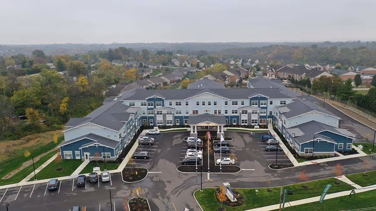 Aerial view of Traditions at North Bend, a large two-story senior living facility with blue and white exterior walls, surrounded by parking lots and green landscaped areas, with a residential neighborhood and trees in the background under an overcast sky.
