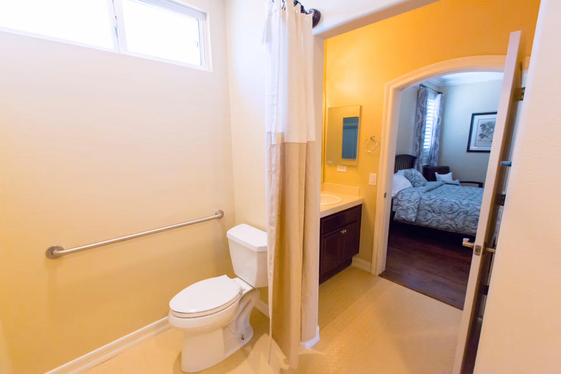 A bathroom with a white toilet, a grab bar on the wall, and a shower curtain partially drawn. There is a window above the toilet letting in natural light. The bathroom opens into a bedroom with a bed covered in a patterned blue and white comforter, a chair, and a framed picture on the wall.