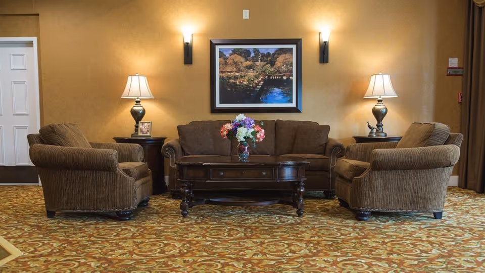A seating area with a brown sofa and two armchairs around a wooden coffee table with a vase of flowers, lamps and framed artwork on the wall.